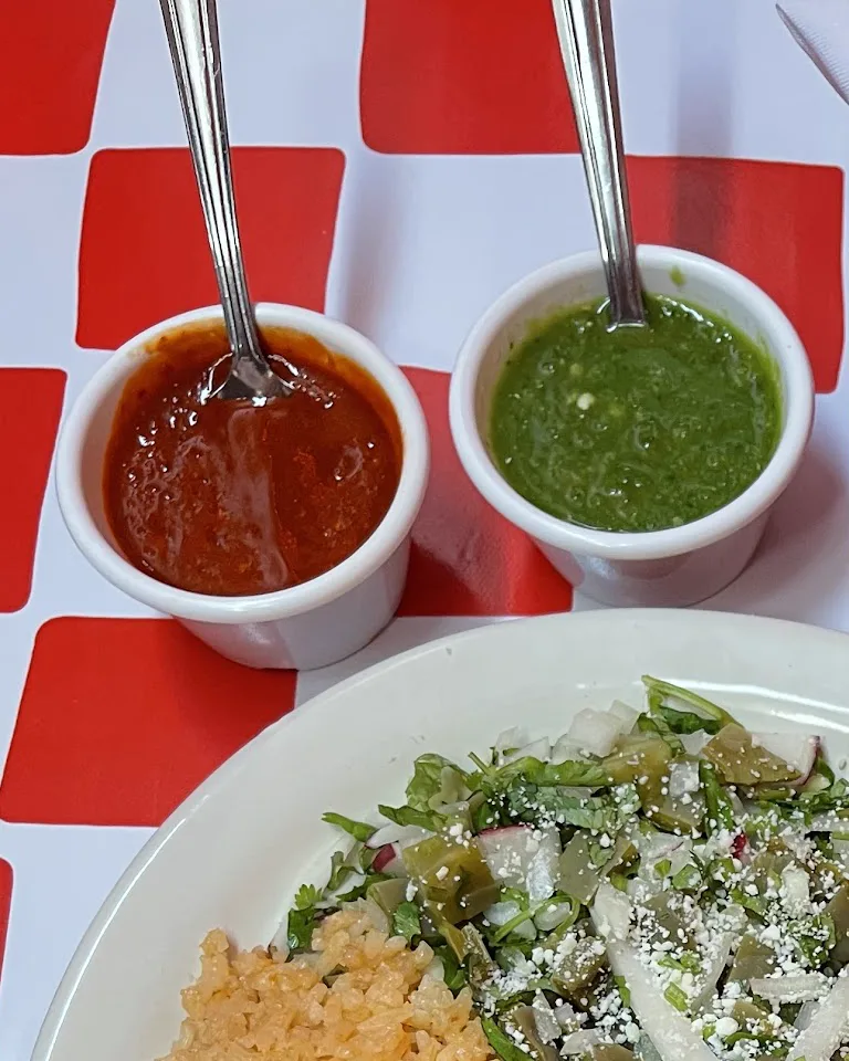 Two small cups of salsa with spoons, one red and one green, on a red and white checkered cloth